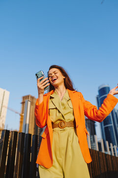 Happy young woman in orange blazer and green jumpsuit laughs while looking at her phone against city skyline. Carefree weekend mood, casual urban lifestyle concept.