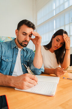 Two people sit across a wooden table, one writes in a notebook while the other leans forward with a thoughtful gaze, conveying collaboration and tension during a study task