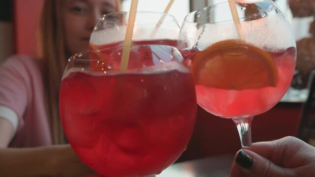 Young woman holds large glass of red beverage with ice and straw another woman hand also holding similar drinks is visible. setting appears to be indoor cafe or bar near windows with soft