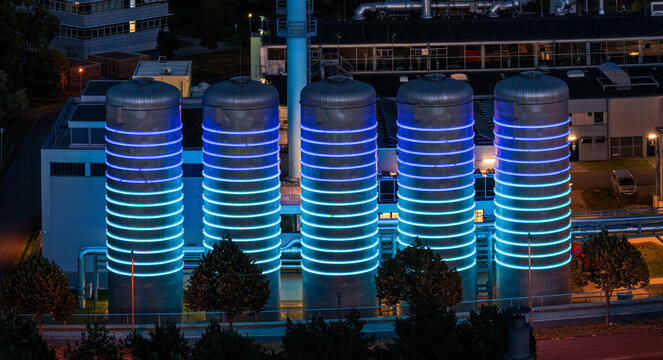 Aerial view of the BTB Heat and Power Plant Adlershof at night, featuring large industrial tanks illuminated with glowing blue LED rings in Berlin, Berlin, Germany.