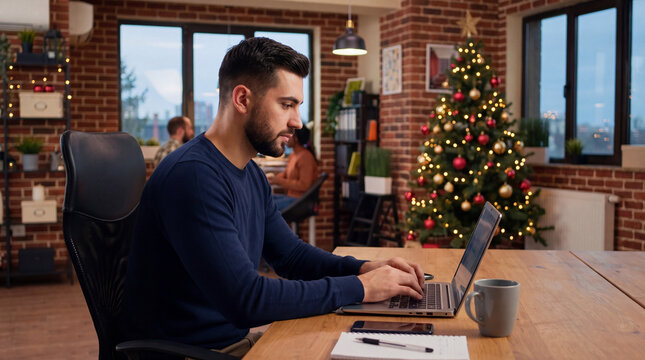 Man working on laptop at wooden table beside decorated Christmas tree in brick interior with winter light through windows
