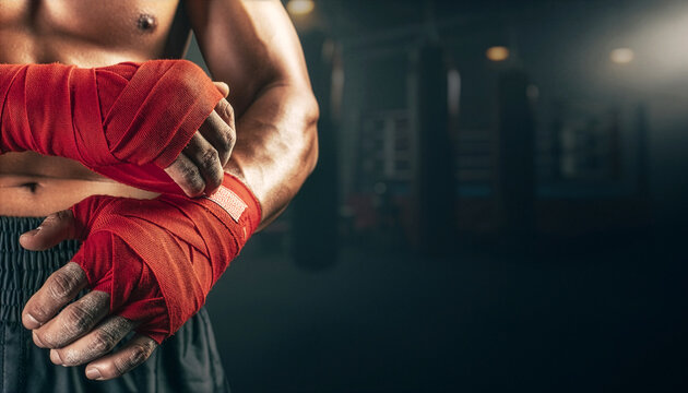 A muscular boxer secures the end of his red hand wraps, preparing inside a dark training gym.