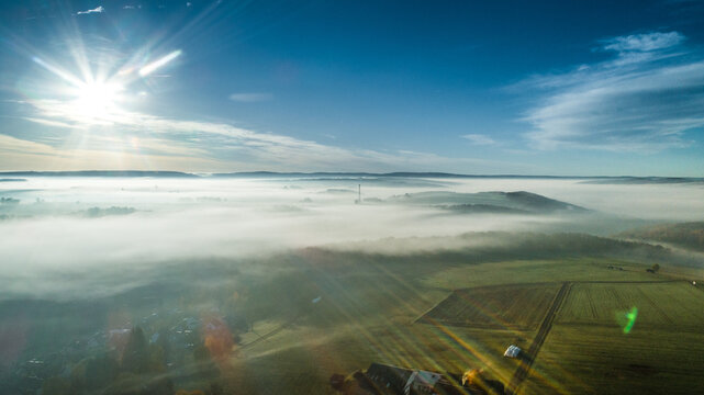 Aerial view of a rural landscape covered in a thick layer of morning fog, with sun rays breaking through the sky and illuminating the fields and trees in Germany.