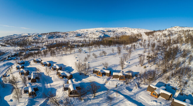 Aerial view of snow-covered cabins nestled in a winter landscape with bare trees and bright sunlight under a clear blue sky in Kirkenes, Finnmark, Norway.