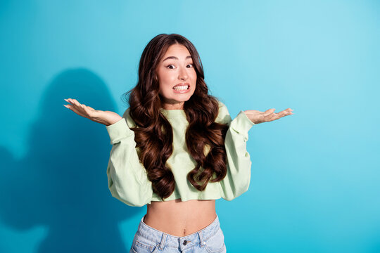 Young woman shrugging with uncertain expression in a casual cropped sweatshirt and jeans against a bright blue background