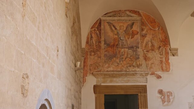 Slow tilt down of a faded medieval fresco of St. Michael above a door in Montescaglioso Abbey, Matera. Peeling plaster highlights the historic texture of the ancient walls.