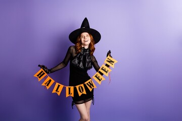 Obraz premium Young woman in witch costume holding Halloween banner against purple background, smiling and celebrating seasonal costume party
