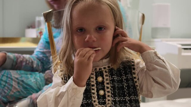 Young girl with blonde hair wearing black and white tweed vest over white blouse eats small piece of food while sitting next to adult.