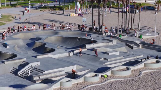 Aerial view displays people skateboarding on concrete bowls and ledges at Venice Beach Skatepark in Los Angeles, California, with spectators gathered on the beach promenade under sunny daylight.