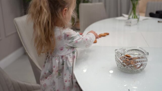 Young blonde girl in floral dress sits at white table, reaching for cookies from glass bowl and arranging them in line.