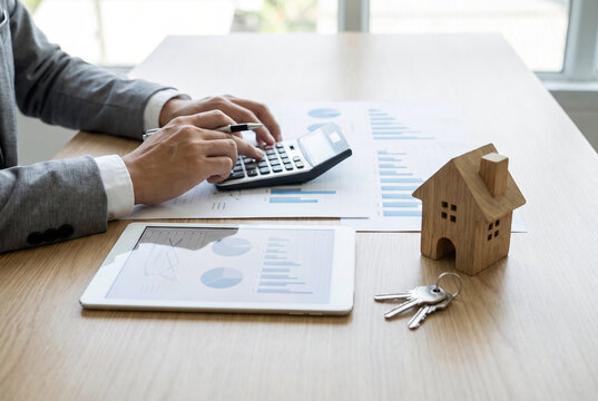 A person uses a calculator at a desk with a tablet displaying charts, a small wooden house, and keys, indicating financial planning and analysis for a property investment.