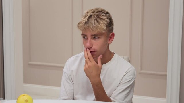 Young blonde-haired boy with his finger to his lips, looking thoughtful while sitting at table with yellow apple.