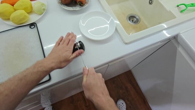 First-person view of a man sharpening a knife using a manual sharpener on a white kitchen countertop, preparing utensils before cooking fresh vegetables like potatoes and carrots.