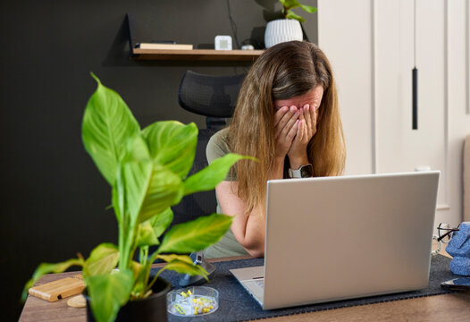 Woman sitting at desk with face covered by both hands in front of open laptop