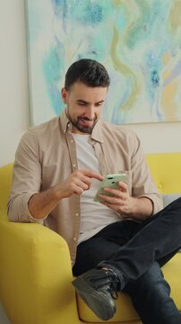 A man relaxes on a bright chair and looks at his mobile device inside a sunlit room with an expressive abstract painting above. The frame captures calm focus and casual digital activity.