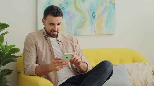 A man uses a mobile phone while lounging on a vivid yellow sofa. The room features a soft neutral wall and a swirling blue teal artwork, creating a relaxed contemporary vibe perfect for tech and