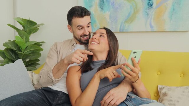 A smiling couple relaxes on a yellow couch as they capture a playful selfie. Warm lighting and soft decor frame a happy moment of togetherness and digital sharing, indoors with green plant accents.