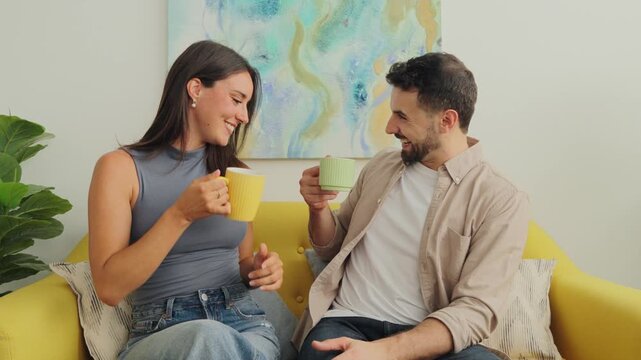 Candid daytime shot of two adults enjoying light conversation while holding mugs on a yellow sofa. Bright wall art and soft natural light create a cheerful relaxed vibe perfect for lifestyle and