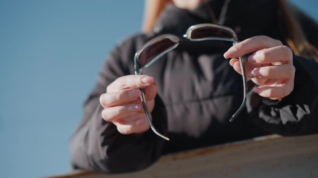 Woman holds and examines pair of sunglasses against bright outdoor light focusing on object in her hands. Her black jacket is partially visible as woman manipulates eyewear with clear blue