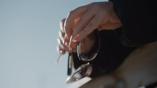 Woman carefully handles pair of sunglasses adjusting frame and inspecting details against outdoor sky backdrop. Her hands are prominently featured as woman examines or prepares to wear