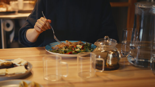 Medium shot of a woman eating dinner with a fork from a blue plate with poultry meat, buckwheat, and a fresh salad of lettuce, red bell peppers, and cucumbers on a wooden table in a cozy warm kitchen.