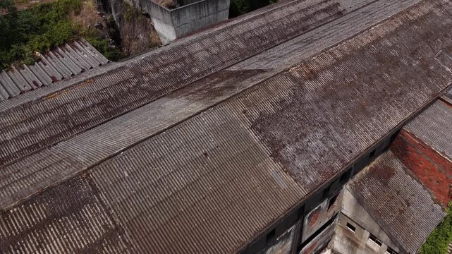 Aerial footage shows a deteriorating asbestos roof atop an abandoned factory, revealing aged corrugated sheets, rust stains, overgrown greenery, and weathered industrial structures.