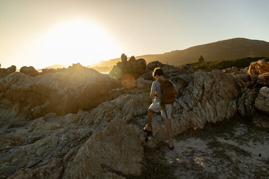 13 year old boy wearing backpack exploring the iconic rocky coastline of Kammabaai Beach, Hermanus, South Africa at sunset