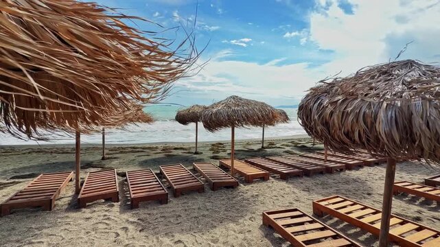Straw umbrellas stand on a sandy beach with wooden lounge chairs arranged in rows. The ocean is in the background, and clouds fill the sky above.