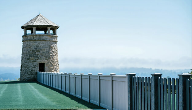 Stone watchtower with a shingled roof next to a long perimeter fence. Security tower on a green lawn with a scenic background