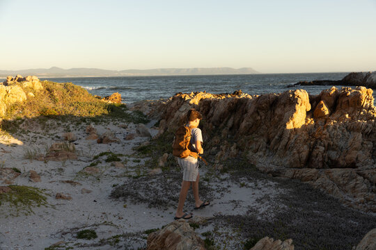 13 year old boy wearing backpack exploring the iconic rocky coastline of Kammabaai Beach, Hermanus, South Africa at sunset