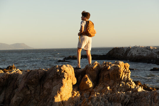 13 year old boy wearing backpack exploring the iconic rocky coastline of Kammabaai Beach, Hermanus, South Africa at sunset