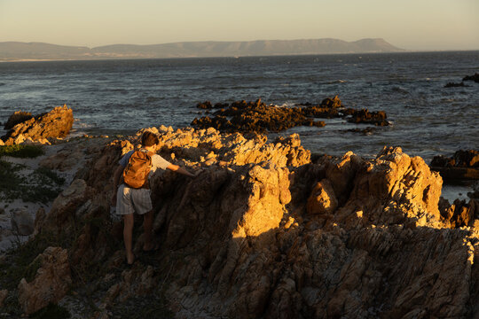 13 year old boy wearing backpack exploring the iconic rocky coastline of Kammabaai Beach, Hermanus, South Africa at sunset