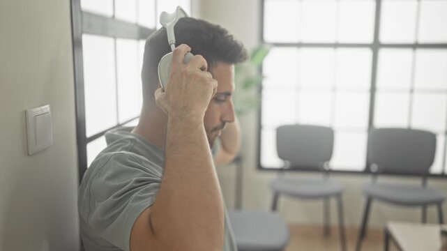 Man wearing headphones sits in a chair with eyes closed, leaning against a wall in a bright waiting area of a building while tapping a red wristband; calm listening.
