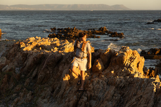 13 year old boy wearing backpack exploring the iconic rocky coastline of Kammabaai Beach, Hermanus, South Africa at sunset