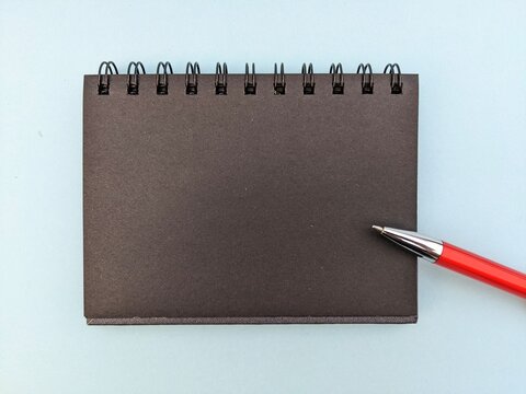 notebook and pen. A top-down view of a blank black spiral notebook and a red ballpoint pen placed on a clean light blue pastel surface. Copy space for text.