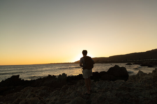 13 year old boy wearing backpack exploring the iconic rocky coastline of Kammabaai Beach, Hermanus, South Africa at sunset