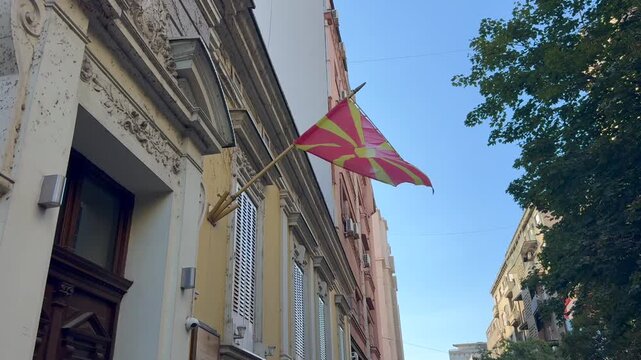 Colorful flag of North Macedonia country hanging from ornate historic building facade on sunny urban street with leafy trees and apartment architecture in a European city