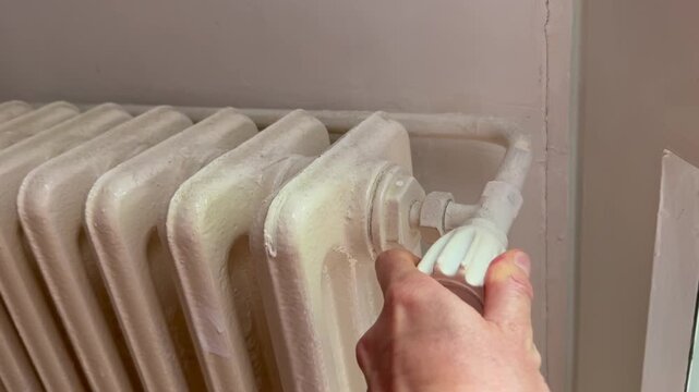 Hand turning a white radiator valve to adjust indoor heating beside a wall, close up view of household maintenance and temperature control