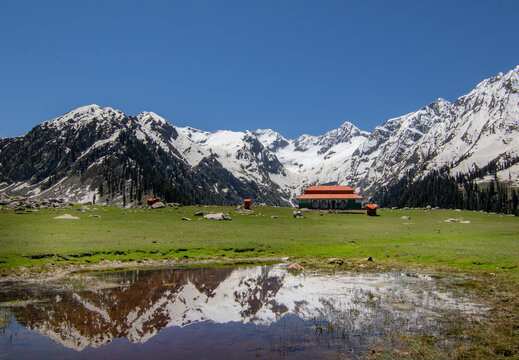 View of Jahaz Banda Alpine Rest House in a green meadow with a pond reflecting snow-capped peaks in Kumrat Valley, Khyber Pakhtunkhwa, Pakistan.