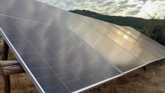 Solar panels reflecting warm morning sunlight across a rural hillside installation surrounded by dry grass, trees, and distant mountains in a renewable energy setting, Green and sustainable energy.