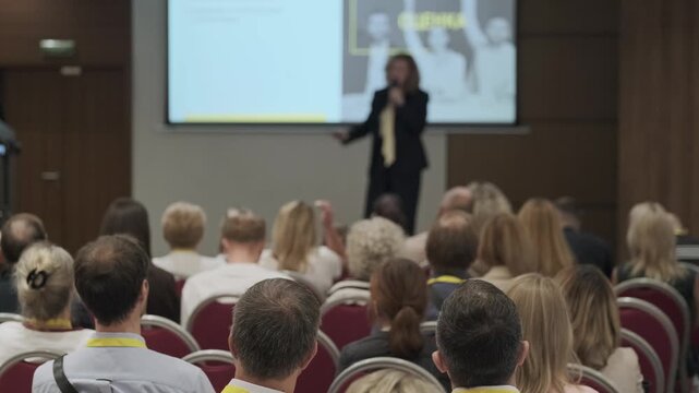 Conference audience focused on presenter during business presentation, networking atmosphere, professional seminar setting.
