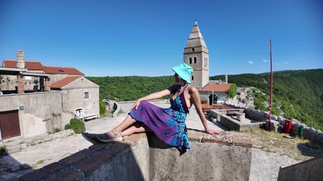 Woman in a colorful dress and hat relaxing on a stone wall. Enjoying the panoramic view of the historic Lubenice village. A medieval settlement on Cres Island. Croatia of Cres Island in Croatia