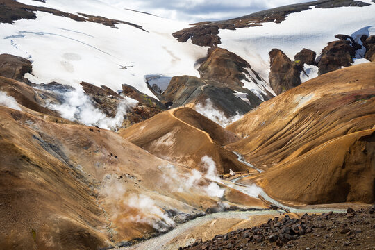 View of Kerlingarfjoll geothermal area with steaming vents, orange rhyolite mountains, snow patches, and a winding hiking trail in Hrunamannahreppur, Iceland.