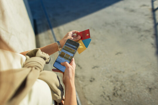 A woman uses a banking app on her smartphone while holding several colored credit cards, facilitating modern mobile payments outdoors in sunny daylight on a concrete surface. E-commerce, creative idea