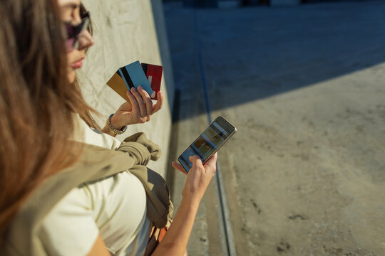Stylish woman holding several colorful credit cards while using a mobile banking application on her smartphone for a cashless online purchase outdoors on a sunny day.