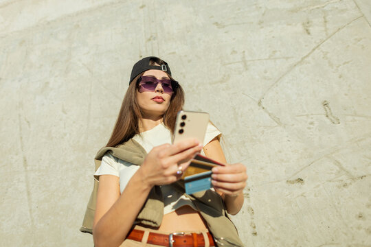 Attractive urban woman in a baseball cap and sunglasses holds a smartphone and a stack of credit cards. She is using mobile banking. Low angle shot against a concrete background.