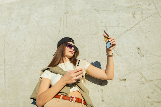 Young trendy woman holding smartphone and multiple credit cards against concrete wall background outdoors. Modern girl selecting payment options for online shopping or mobile banking app.