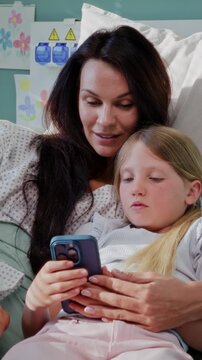 Mother and daughter use smartphone lying together on hospital bed
