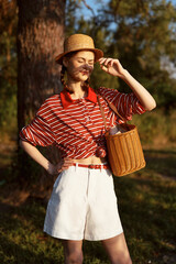 Obraz premium Young woman in a straw hat and striped shirt with a wicker bag poses outdoors during golden hour sunlight, enjoying a summer lifestyle while standing relaxed in a sunlit forest glade.