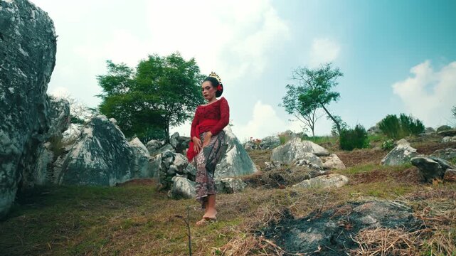 A female model showcasing traditional Indonesian fashion, standing elegantly with one hand on her waist in a natural rocky environment.